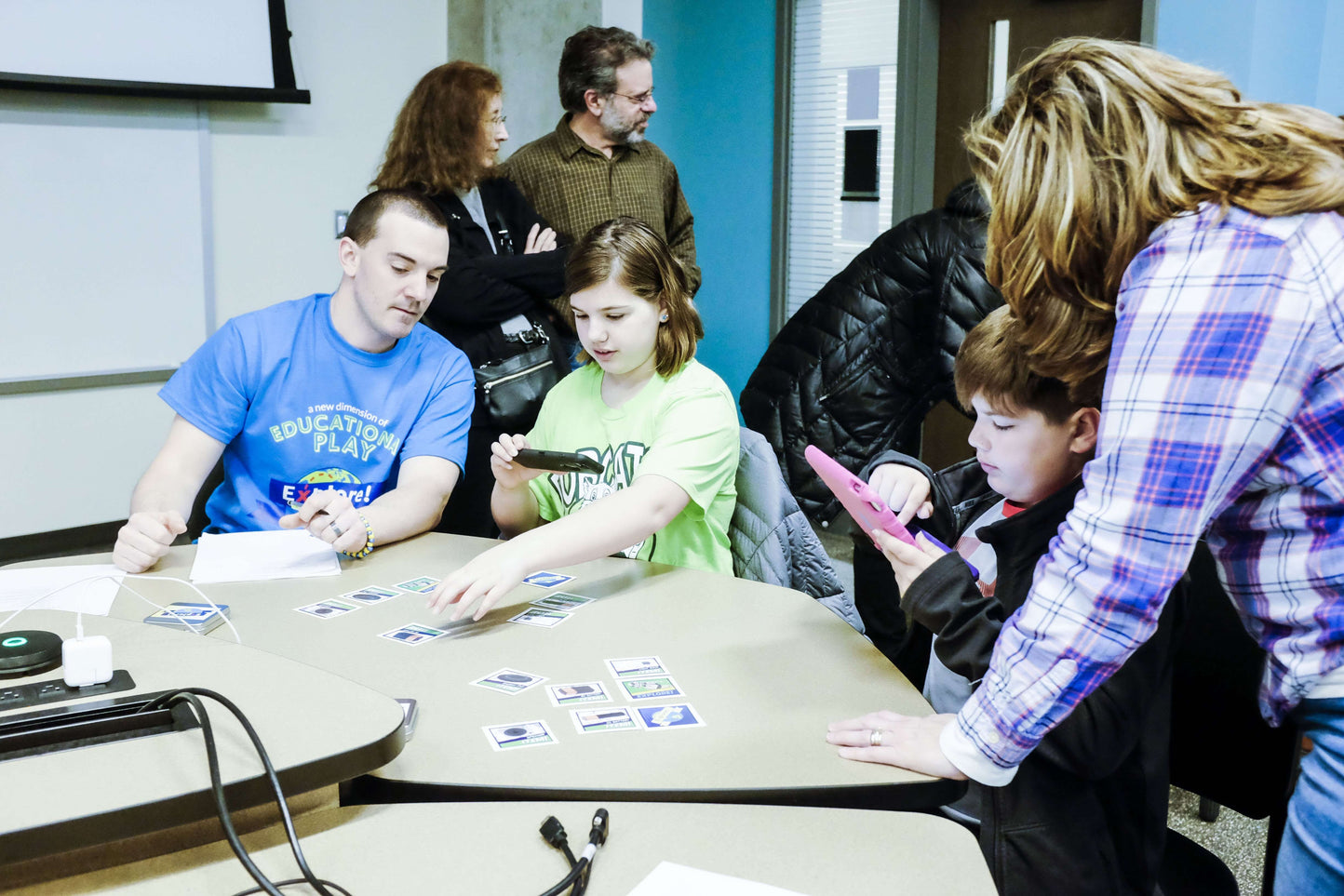 Group of people, including children and adults, engaged in an augmented reality educational activity with cards on a table.
