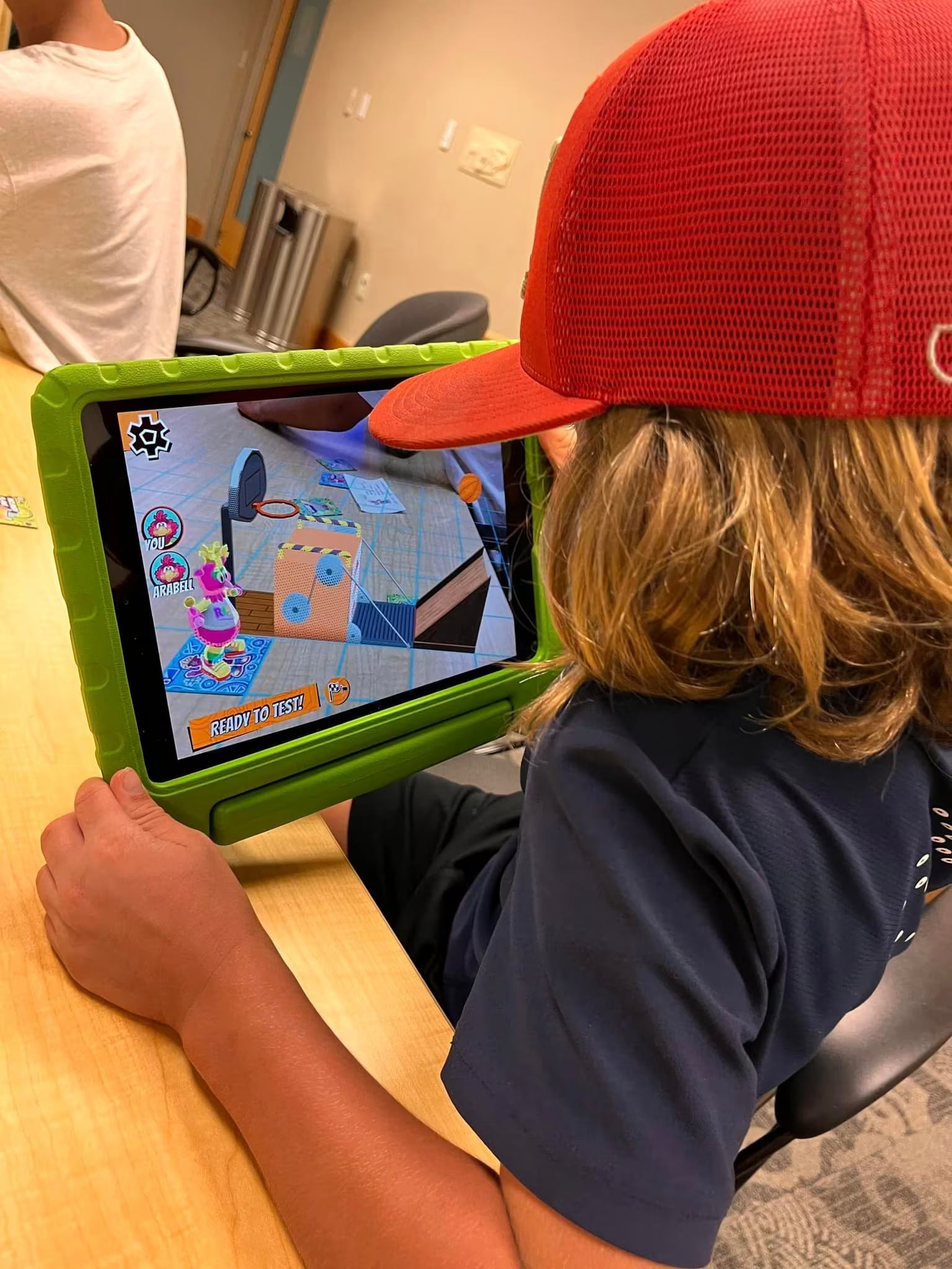 Child wearing a red cap using a tablet with a colorful app on a wooden table.